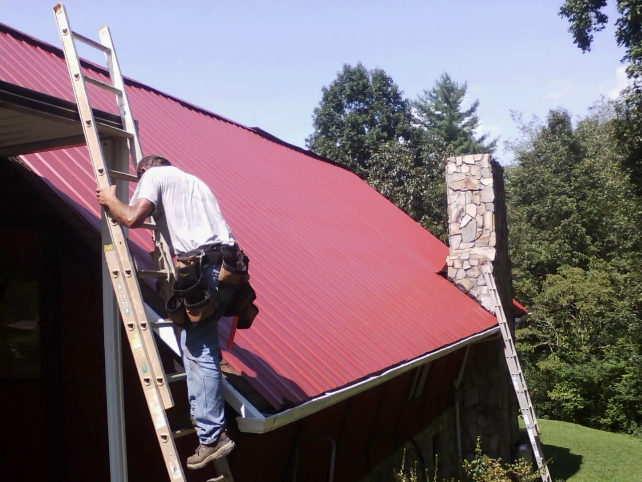 Roofer on a ladder beside a red metal roof and stone chimney — Soffit & Fascia Repair by Roofing Brothers in Hatfield